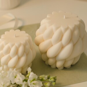 Könnte beinhalten: Zwei cremefarbene, strukturierte Kerzen mit geflochtenem Design. Die Kerzen haben eine blütenartige Oberseite und stehen auf einer hellgrünen Oberfläche, mit weißen Blumen im Vordergrund. Dekorative Wohnaccessoires.