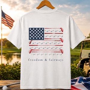 Könnte beinhalten: Weißes T-Shirt mit einem Design der amerikanischen Flagge, das Golfschläger und -bälle enthält. Der Text "freedom & fairways" ist darunter aufgedruckt. Im Hintergrund sind ein Golfplatz und die amerikanische Flagge zu sehen.