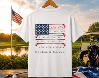 Könnte beinhalten: Weißes T-Shirt mit einem Design der amerikanischen Flagge, das Golfschläger und -bälle enthält. Der Text "freedom & fairways" ist darunter aufgedruckt. Im Hintergrund sind ein Golfplatz und die amerikanische Flagge zu sehen.