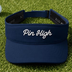 May include: A navy blue golf visor featuring the white embroidered text Pin High on the front panel. The visor has an adjustable strap at the back with a black buckle and interior branding that reads 110 FLEXFIT.