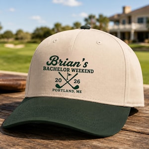 May include: A beige baseball cap with a dark green brim featuring dark green text that reads Brian's BACHELOR WEEKEND 20 26 PORTLAND, ME with a graphic of two crossed golf clubs and a flag centered on the front panel of the hat.