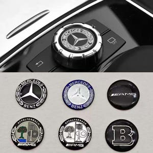 Può includere: Una collezione di emblemi per auto Mercedes-Benz e una manopola di controllo. Gli emblemi includono il logo Mercedes-Benz, AMG e Brabus. La manopola di controllo è argentata e nera con il logo Mercedes-Benz.