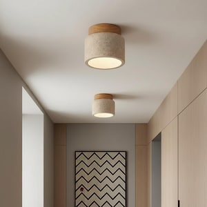 May include: Two cylindrical ceiling lights with a light beige speckled body and a wooden top. The lights are mounted on a white ceiling in a hallway. The lights are turned on, emitting a warm glow.