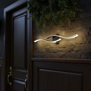 Puede incluir: Lámpara de pared exterior moderna con un diseño fluido en forma de ola. La lámpara está hecha de metal oscuro y presenta dos brazos curvos e iluminados que emiten una luz blanca cálida. Está montada en una pared de piedra oscura.