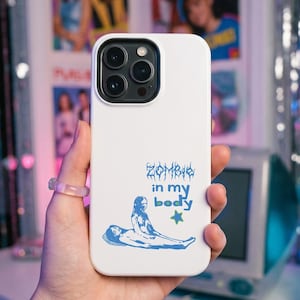 May include: A white smartphone case featuring a blue graphic illustration of two figures and the text "ZOMBIE in my body" written in a stylized font, accompanied by a small green star icon, with a black camera lens cutout at the top.