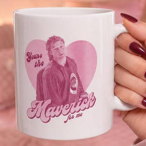 Puede incluir: Taza de cerámica blanca con un diseño de corazón rosa, una imagen de hombre y el texto "You're the Maverick for me" en rosa. La taza tiene un asa curva y una superficie lisa.