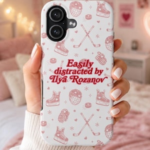 May include: A white phone case featuring a pink hockey-themed pattern with skates, helmets, pucks, and crossed hockey sticks. The text "Easily distracted by Ilya Rozanov" is in pink. The case is held in a person's hand.