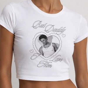 May include: White crop top with a black and white graphic of a man inside a heart-shaped frame. The text "Bad Daddy I Love Him" is above and below the image.
