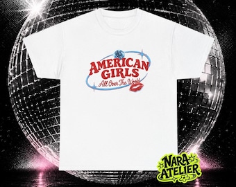 May include: A white short-sleeve t-shirt featuring a graphic print with the red text AMERICAN GIRLS and the script text All Over The World in blue, accompanied by a small blue disco ball, four-pointed stars, and a red lipstick kiss mark.
