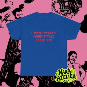 Puede incluir: Una camiseta de manga corta en color azul real con el texto en rojo I SUPPORT MY WIFE'S HARRY STYLES ADDICTION impreso en una fuente pixelada en el centro del pecho. La prenda tiene cuello redondo y un corte recto estándar.
