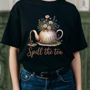 May include: Black t-shirt featuring a floral teapot design with the text "Spill the tea". The teapot is filled with daisies and other flowers, with tea pouring out. The design is in watercolor style.