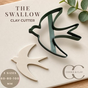Könnte beinhalten: Ein dunkelgrüner Ton-Ausstecher in Schwalbenform und ein passendes, cremefarbenes Ton-Exemplar. Der Text lautet THE SWALLOW CLAY CUTTER, 3 SIZES 60-80-100 MM und COOKIE & CLAY. Der Ausstecher zeigt die Silhouette eines Vogels mit spitzen Flügeln.