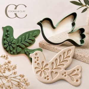 Pode incluir: Três objetos em forma de pássaro, incluindo um carimbo verde-escuro com detalhes florais e de coração, um carimbo bege com padrões em relevo correspondentes e um cortador de contorno verde-escuro. O texto COOKIE & CLAY é visível no canto superior esquerdo.