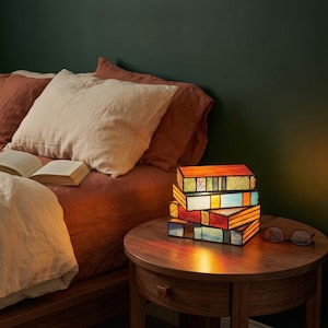 May include: A colourful stained glass lamp in the shape of stacked books, with orange, red, blue, and green panels. The lamp is on a wooden bedside table next to a bed with rust-coloured bedding. A pair of glasses rests on the table.
