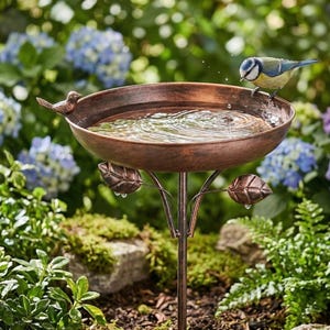 May include: A bronze-colored bird bath with a small bird perched on the edge. The bath is filled with water, and the stand has leaf-shaped accents. The background features lush greenery and blue flowers, creating a natural garden setting.