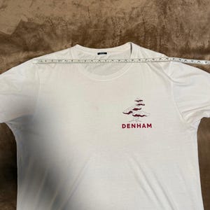 Puede incluir: Camiseta blanca de cuello redondo con un gráfico de árbol granate y el nombre de la marca "DENHAM" impreso debajo. La camiseta es de un material suave y se mide con una regla.