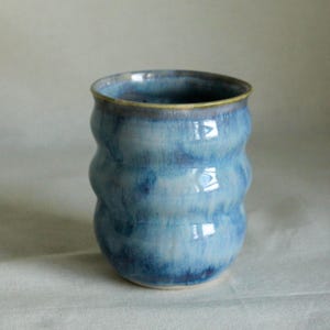 May include: A ceramic tumbler featuring a wavy, rippled silhouette and a glossy glaze in varying shades of blue. The surface displays a blended, mottled pattern of light and dark azure tones with a smooth, rounded rim and a solid, stable base.