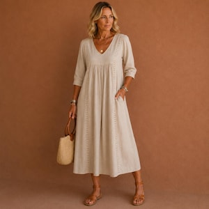 May include: A beige midi dress with a V-neck, three-quarter sleeves, and pockets. The dress features decorative stitching down the front and is paired with brown sandals and a straw tote bag. The woman is wearing a gold necklace and bracelets.