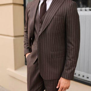 May include: A three-piece brown suit with white pinstripes. The suit includes a blazer, vest, and trousers. The suit is paired with a white dress shirt and a brown tie. The text "Tailored Tales Crafts" is at the top of the image.