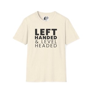 Puede incluir: Camiseta color crema con el texto "LEFT HANDED & LEVEL HEADED" en letras negras. La camiseta tiene cuello redondo y mangas cortas. La tela es texturizada.