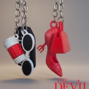 May include: Silver chain necklaces with charms. One necklace features a coffee cup, sunglasses, and a black rectangle. The other necklace has a red high heel shoe and a red purse. The text "THE DEVIL WEARS PRADA" is at the bottom.