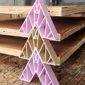 May include: Three geometric, triangular shelves in shades of pink and beige are stacked in a tiered arrangement. Each shelf features a lattice design and is secured with a small, silver-colored magnet. The shelves are set against a backdrop of wooden planks.