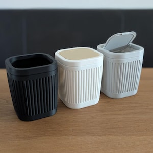 May include: A white ceramic mug with a black handle and the text "But First, Coffee" in a black, handwritten-style font. The mug is on a white surface.