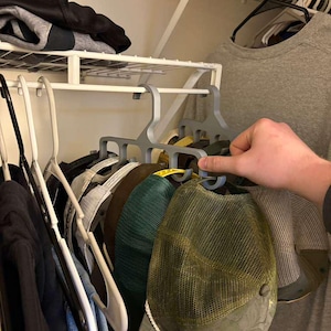 May include: A gray hat rack holding several baseball caps of various colors and styles. The caps include a green mesh trucker hat, a brown cap, and others. The rack is in a closet with other clothing items.