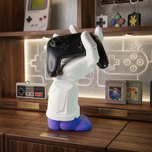 May include: A white, stylized hand-shaped stand with a blue base, holding a black wireless video game controller. The hand features a segmented, robotic design with a cuff at the wrist, providing a stable mount for the controller in a vertical position.