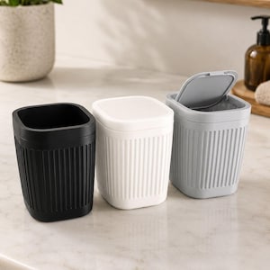 May include: Three small, square-shaped waste bins with a vertical ribbed texture on the exterior. One bin is matte black, one is solid white, and the third is light gray with a hinged lid that is partially open, revealing the interior compartment.