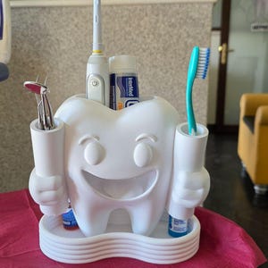 May include: A white, tooth-shaped organizer holding dental tools and supplies. It features a smiling face and two cylindrical holders on either side. An electric toothbrush, toothpaste, and a manual toothbrush are visible. The organizer sits on a tray.