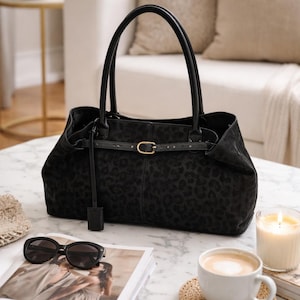 May include: A black handbag featuring a subtle tonal leopard print pattern, two top handles, and a horizontal strap across the front secured with a gold-toned metal buckle. The bag has a structured base and a smooth, matte finish throughout its exterior.