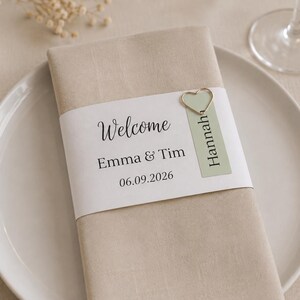 Könnte beinhalten: Eine beige Stoffserviette, umwickelt mit einer weißen Papierbanderole, die den gedruckten Text Welcome, Emma & Tim sowie 06.09.2026 trägt. Ein hellgrünes Namensschild mit dem Namen Hannah ist mit einem silbernen herzförmigen Clip befestigt.