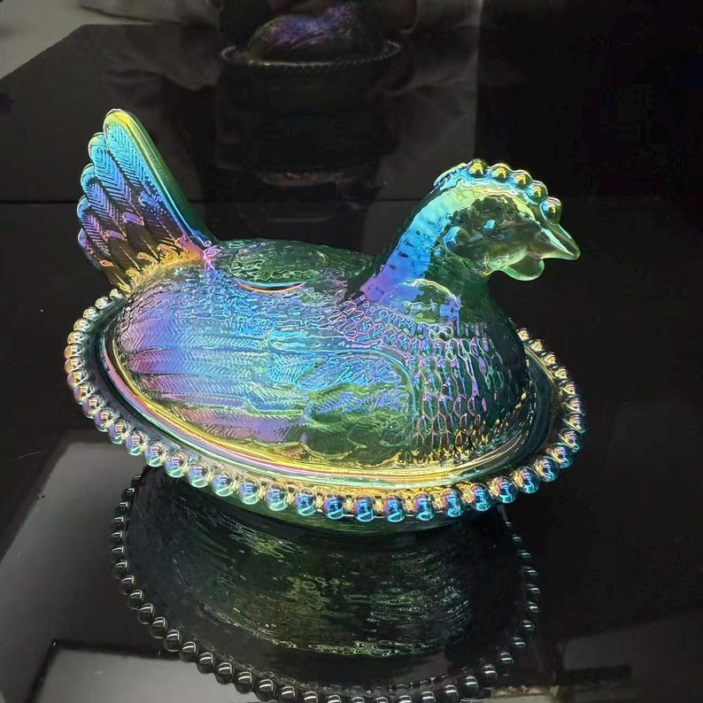 May include: Iridescent glass hen-shaped dish with a lid. The dish is a vibrant mix of blues, greens, and purples. The hen is sitting on a beaded oval base. The lid is shaped like the hen's body and head.