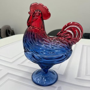 May include: A rooster-shaped glass dish featuring a vibrant color gradient that transitions from a deep red at the comb and head to a rich blue at the base. The piece has a textured, molded surface with detailed feather patterns throughout.