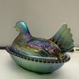 May include: Iridescent glass hen on nest. The hen is a mix of blue, purple, and green, with textured details. The nest is a matching green with a beaded rim. A decorative kitchen or dining room piece.