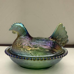 May include: Iridescent glass hen-shaped covered dish. The hen is perched atop a round base with a beaded rim. The glass has a shimmering, multi-colored finish, with hues of blue, green, and purple. The hen's tail feathers are fanned out.