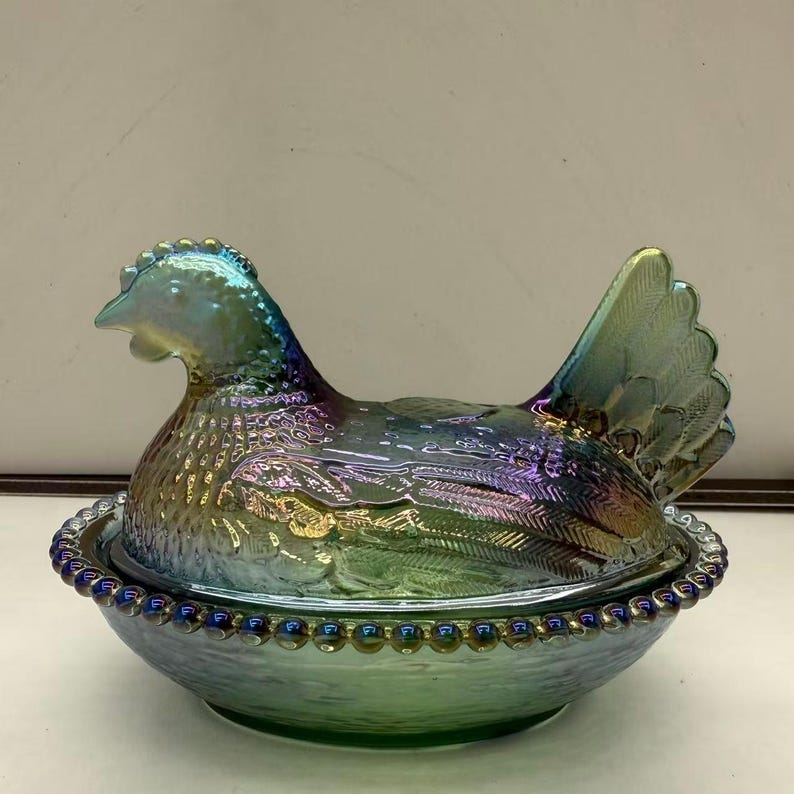 May include: Iridescent glass hen-shaped covered dish. The hen is perched atop a round base with a beaded rim. The glass has a shimmering, multi-colored finish, with hues of blue, green, and purple. The hen's tail feathers are fanned out.