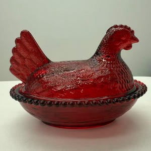 May include: A ruby red glass hen-shaped covered dish. The hen has detailed feather and head features, with a textured surface. The dish sits in a matching base with a beaded rim. A decorative kitchenware item.