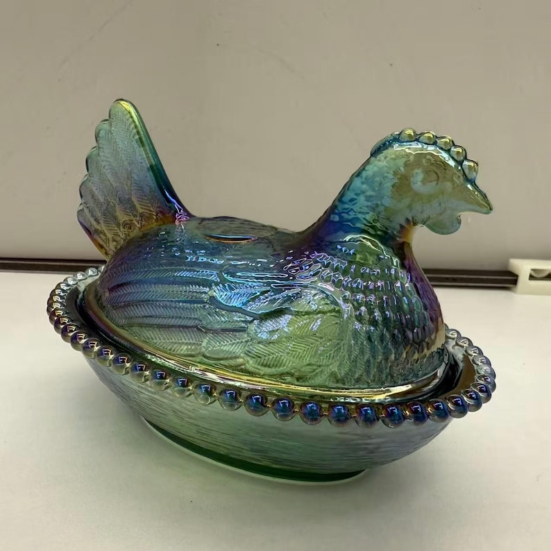 May include: Iridescent glass hen-shaped dish with a lid, sitting in a nest-like base. The dish features a detailed design with a textured surface and a beaded rim. The colors shift from blue to green.