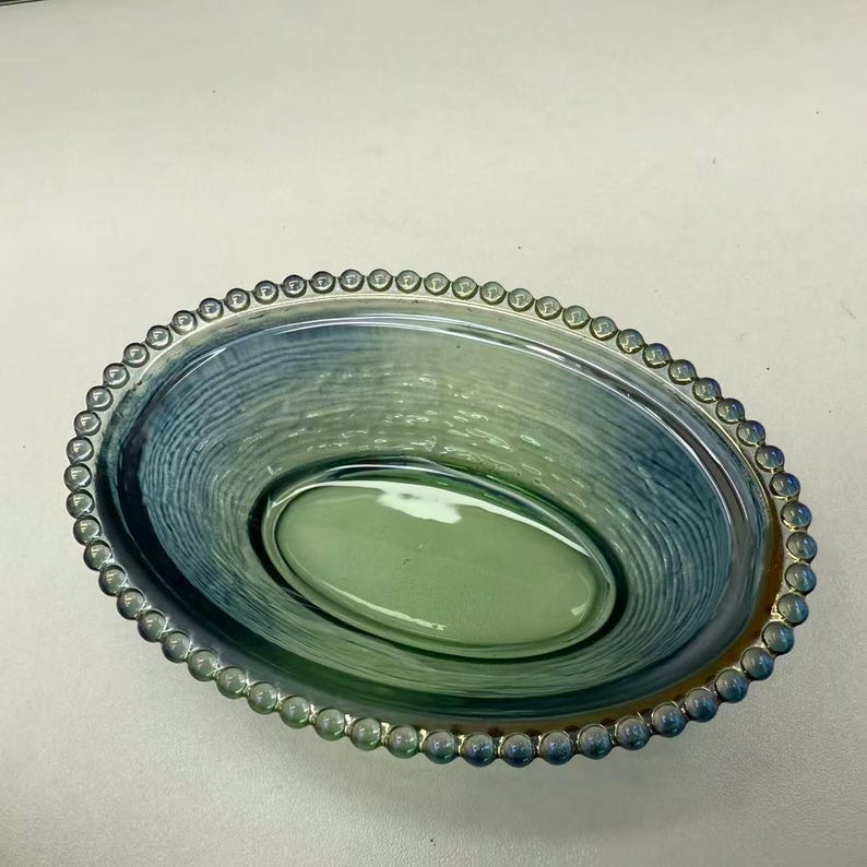 May include: An oval-shaped glass bowl with a textured interior and a beaded rim. The bowl features shades of blue and green, creating a gradient effect. The rim is silver-colored, adding a decorative touch to the piece.