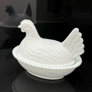 May include: A white milk glass hen on a nest dish featuring a detailed molded chicken lid with textured feather patterns resting on a base with a beaded rim and a woven basket weave design on the exterior surface of the oval-shaped container.