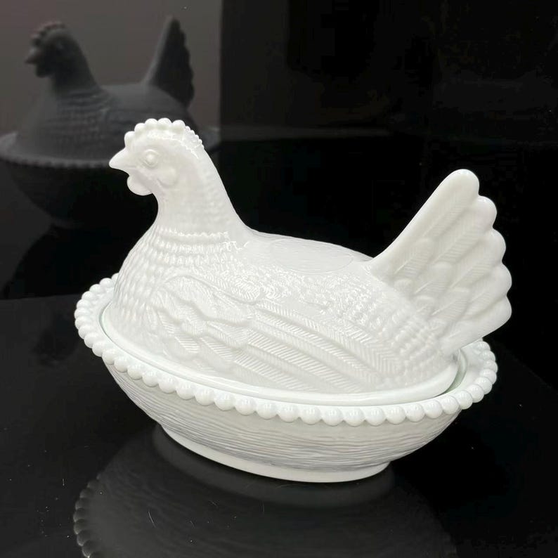 May include: A white milk glass hen on a nest dish featuring a detailed molded chicken lid with textured feather patterns resting on a base with a beaded rim and a woven basket weave design on the exterior surface of the oval-shaped container.