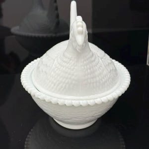 May include: A white milk glass hen on nest dish featuring a detailed chicken lid with textured feather patterns. The base of the dish is shaped like a woven basket and is finished with a decorative beaded rim along the top edge of the bowl.