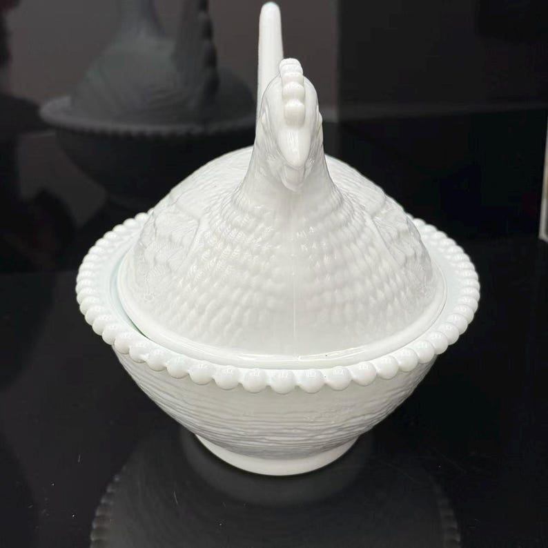 May include: A white milk glass hen on nest dish featuring a detailed chicken lid with textured feather patterns. The base of the dish is shaped like a woven basket and is finished with a decorative beaded rim along the top edge of the bowl.