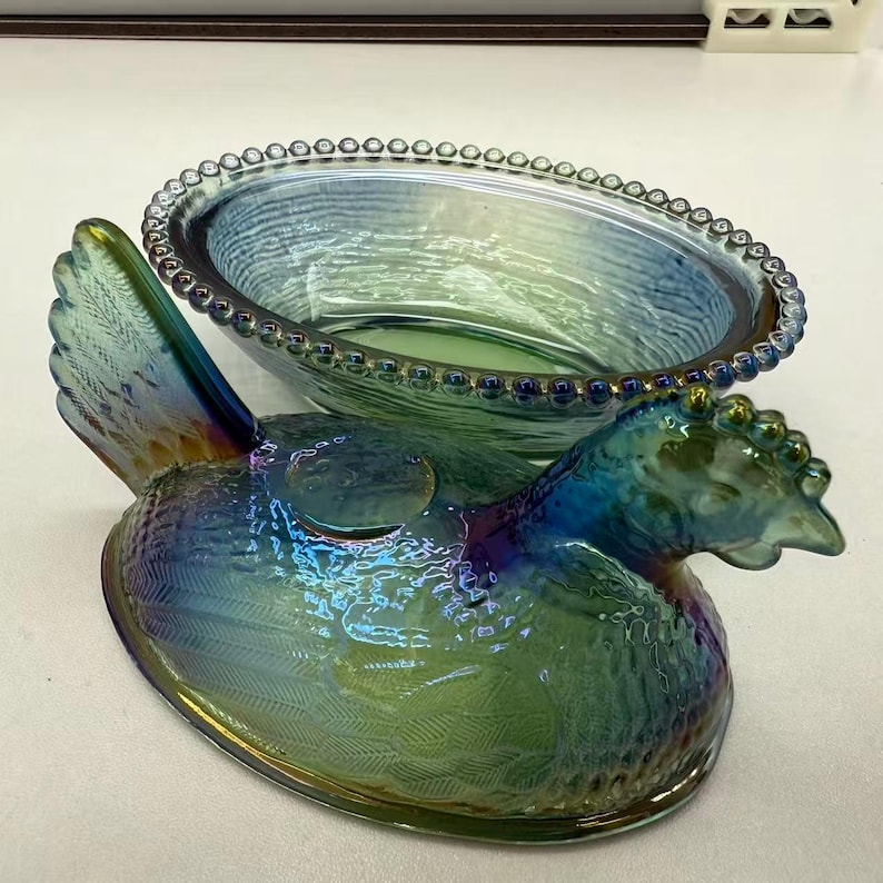 May include: Iridescent glass hen-shaped dish with a matching lid. The dish is primarily green and blue with a beaded rim and feather detailing. The hen's head is raised, and the dish is oval-shaped.