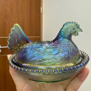 May include: Iridescent glass hen-shaped covered dish. The hen is perched on a nest-shaped base, with a detailed feather design and a beaded rim. The glass has a shimmering, multi-colored finish, reflecting hues of blue, green, and purple.