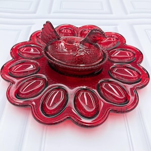 May include: A deep red pressed glass egg plate featuring a central lidded dish shaped like a sitting hen. The circular tray has twelve oval indentations for eggs, with a scalloped edge and a textured pattern throughout the translucent glass surface.