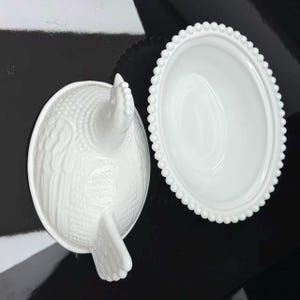 May include: A white milk glass hen on nest dish consisting of two pieces. The lid is shaped like a sitting hen with textured feather details, while the base is an oval bowl featuring a decorative beaded rim along the outer edge of the dish.