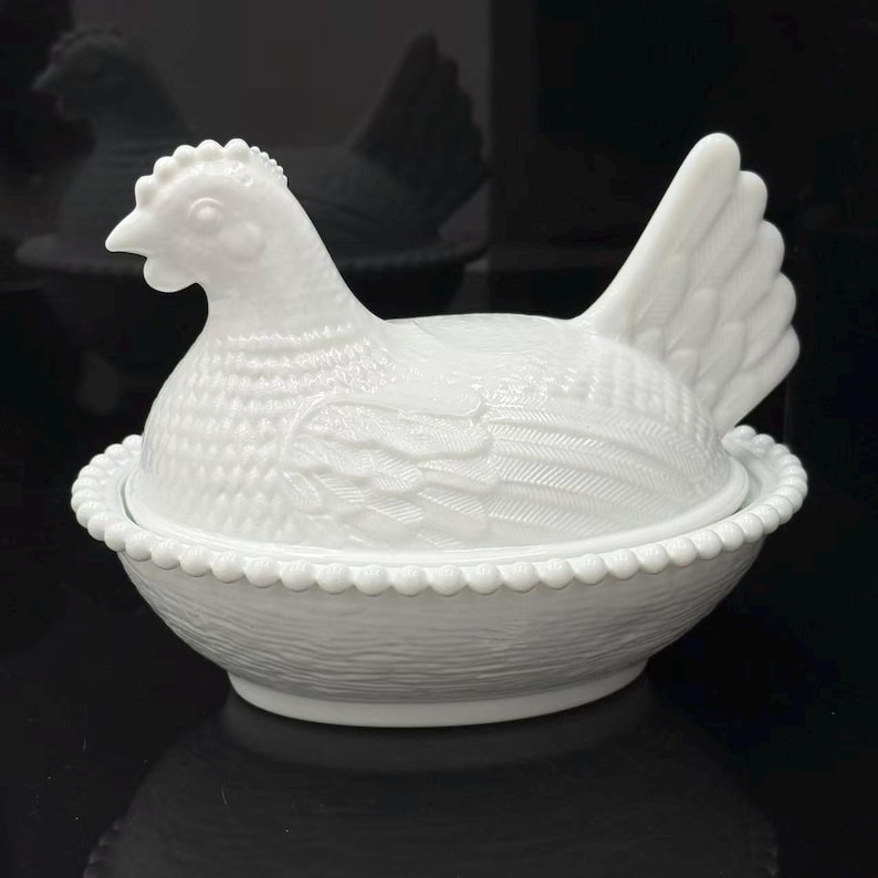 May include: A white milk glass hen on nest dish featuring a detailed chicken lid with textured feathers and a comb, resting on a base with a beaded rim and a woven basket pattern. The opaque glass container is shaped like a sitting hen.